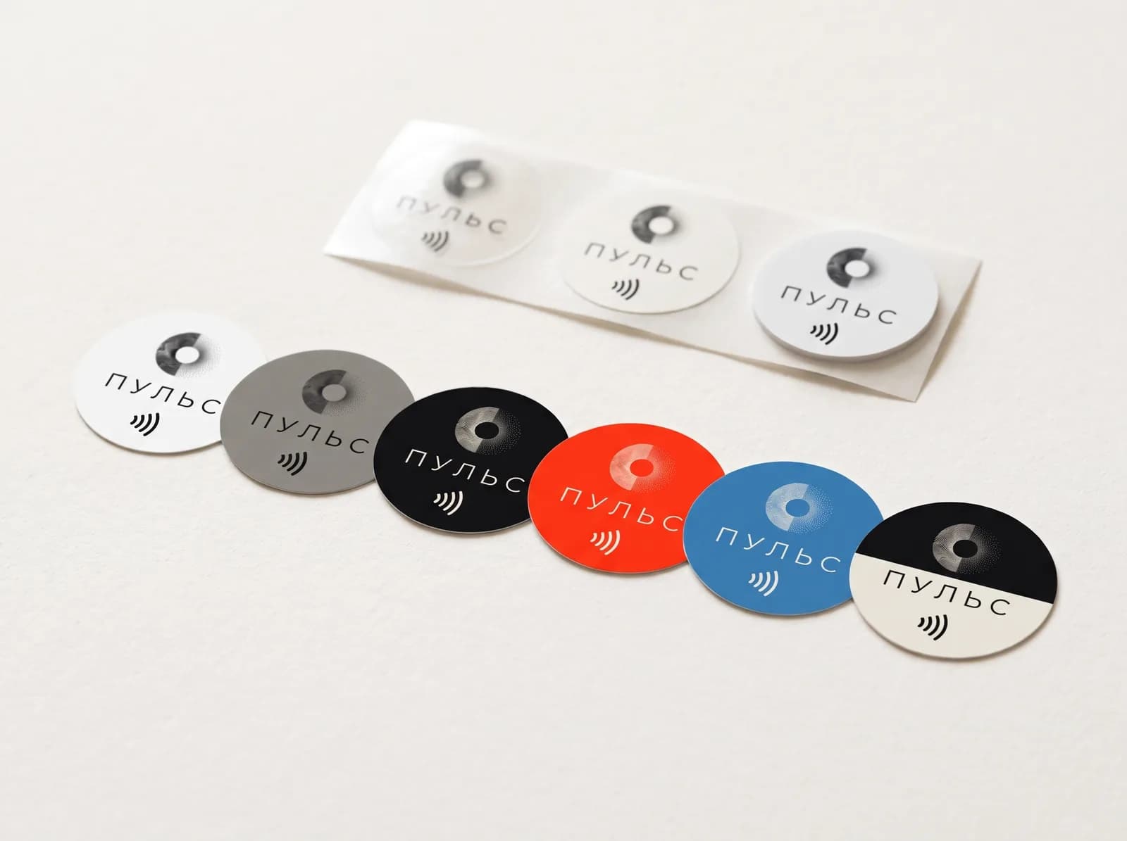 Круглые NFC-наклейки в ряд: белая, серая, чёрная, красная, синяя и со своим макетом, позади три основы на ленте